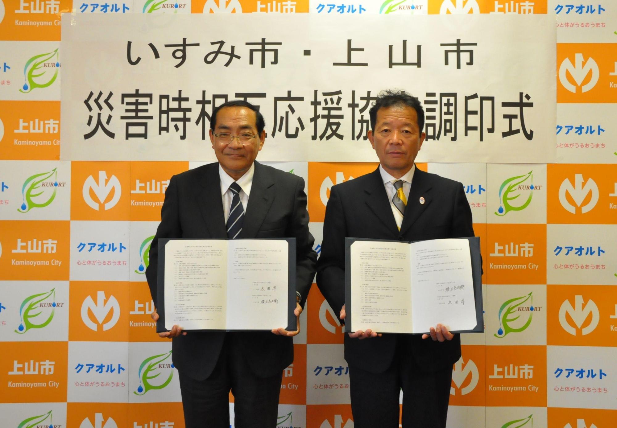「いすみ市・上山市災害時相互応援協定調印式」と書かれた横断幕をバックに、左側にいすみ市長と右側に上山市横戸市長両者が並んでそれぞれが協定書を開いて見せている様子の写真