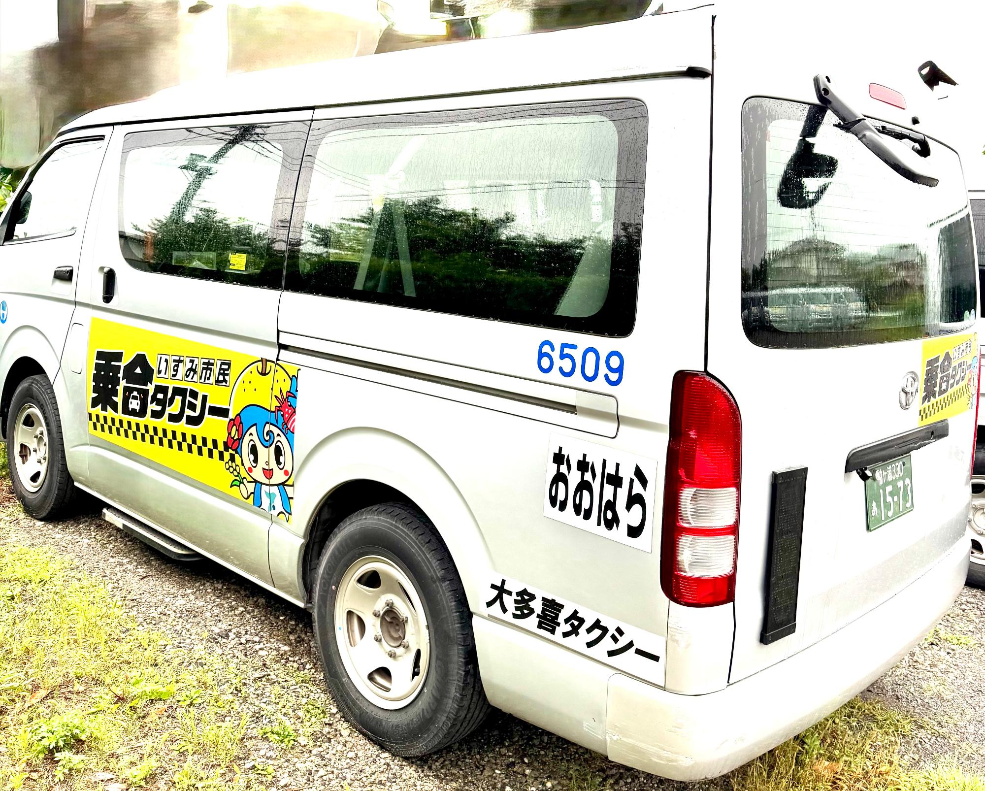 車体の左斜め後ろから撮影し、側面中央付近に黄色の背景に黒字で「いすみ市民乗合タクシー」のロゴがあり、側面後方に上から「6509」「おおはら」「大多喜タクシー」の文字があり、後方のガラスの右下に側面と同じロゴがある写真