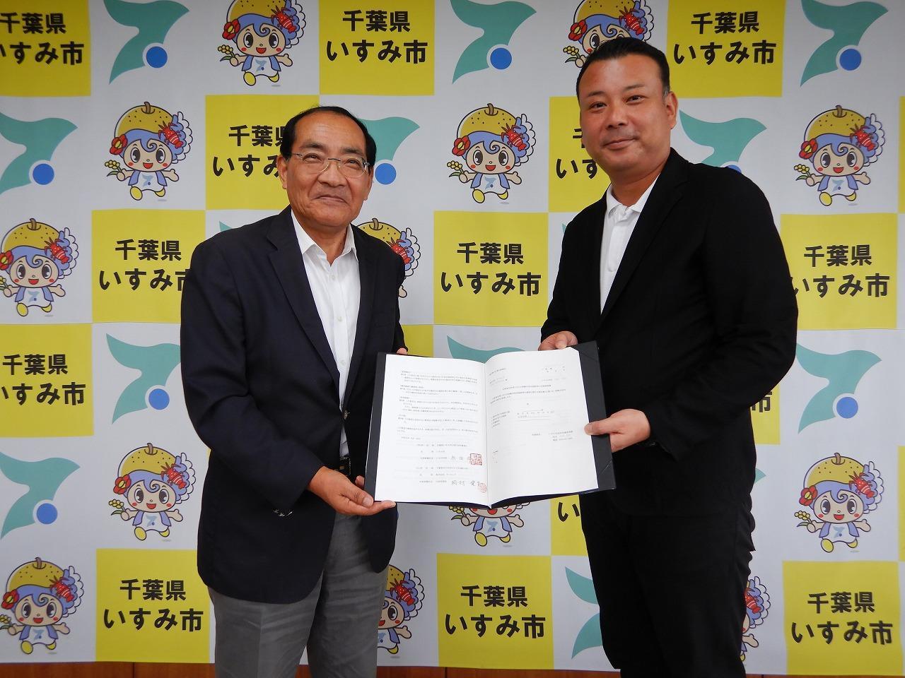 マスコットキャラクターやいすみ市章のカラフルな壁紙の前で、見開きの書類を男性二人が一緒に持ってこちらに笑顔を向けている写真