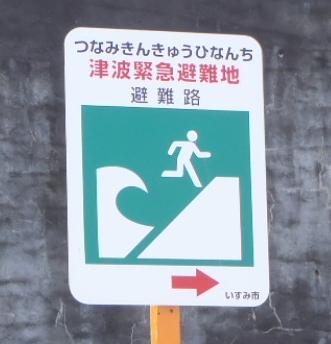 「津波緊急避難地」と赤字で書かれ、避難路の方向を赤の右矢印で示し、人が津波から逃げる様子のイラストが描かれた看板の写真