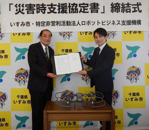 いすみ市のカラフルな壁紙の前に立ち、フェイスシールドマスクを付けた大田市長と伊藤副理事長が見開きの書類を一緒に両手で持ってこちらに見せている写真