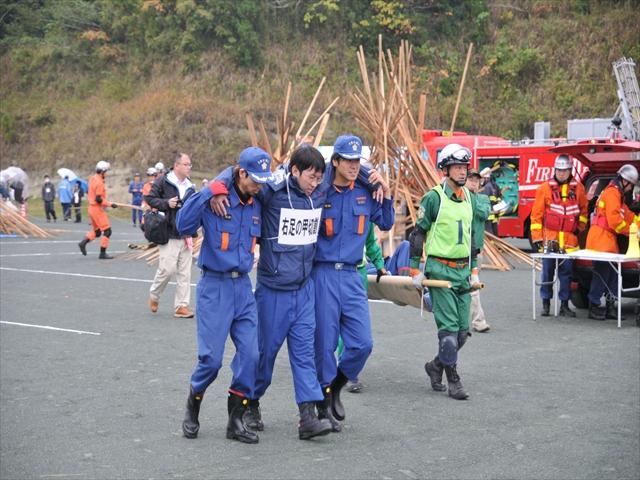 負傷の状態が書かれたパネルを首から下げた男性を両脇から支えるようにして搬送する2名の隊員の写真