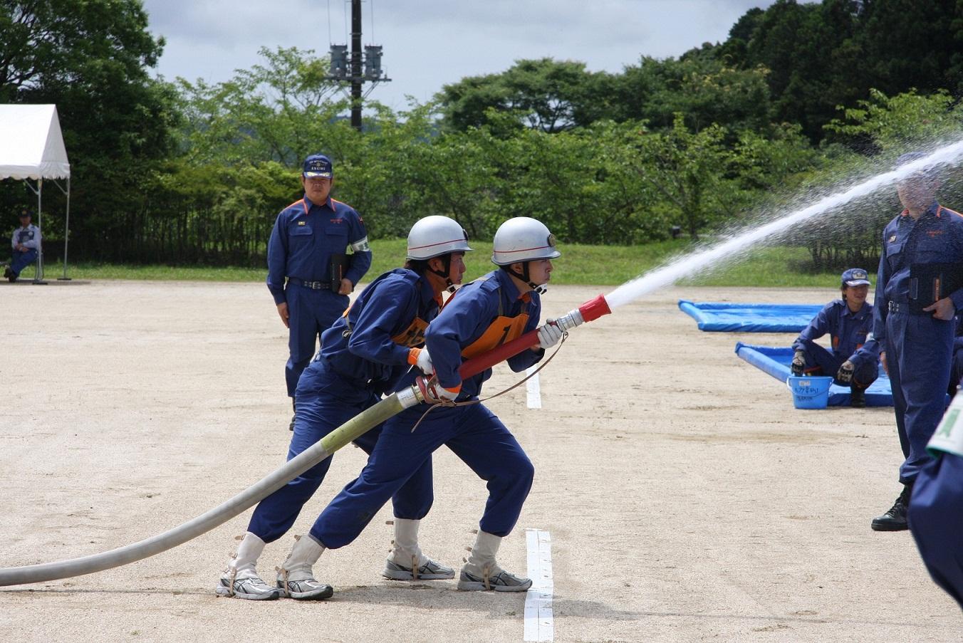 木々にかこまれたグラウンドで、2名の消防団員が前後に並び1本のホースを抱えるようにして持ち、放水している様子を数名が見ている写真