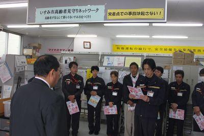 郵便局内で制服の職員が横に並んでそれぞれ書類を手に持写真ち、向かい合って立つスーツ姿の男性の後ろ姿が手前に写る写真