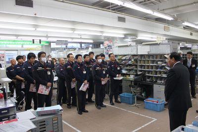 郵便局内で制服を着た職員が書類を手で持ち整列し、向かい合ってスーツ姿の男性が1人立っている写真
