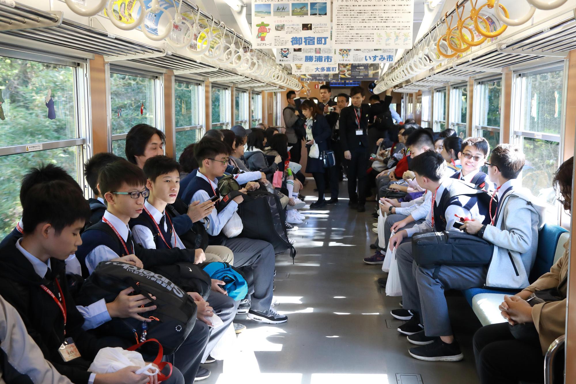 学生の皆さんが、暖かな光が差し込む電車の車両に左右に別れて座りながら談笑する様子の写真