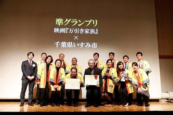 準グランプリ、映画「万引き家族」×千葉県いすみ市と書かれたスクリーンの前に集まって表彰状を掲げながら記念撮影する、黄色い法被を着た人達の写真
