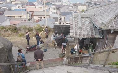 瓦屋根の民家の軒先で機材を手に撮影をしている人たちに様子を、石段の上から見下ろすように捉えた写真