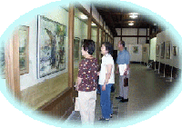 いすみ市郷土資料館内の展示室にて、ガラス張りの展示用ケースの前で展示されている絵画を眺める3人の男女の写真