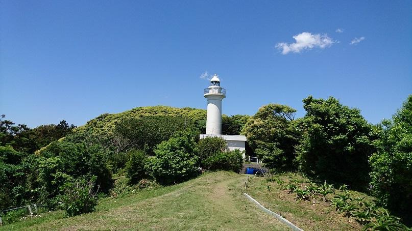 青空の下、小高い所に、木々の緑に囲まれて立つ白い灯台の写真