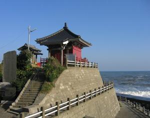 快晴の青空の海岸沿いに高台が有り、階段を登ると、赤い地蔵尊が立てられている写真