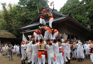 神社で大勢の男性が組体操の3段ピラミッドを作り、最上段で扇子を持ち立っている写真