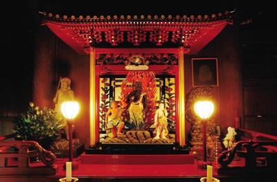 薄暗い室内で、手前の2つの照明の奥に屋根のある赤い豪華な造りのお堂があり、2体の像に挟まれて中央に赤い炎を背にした不動明王像が立っている文化財の写真